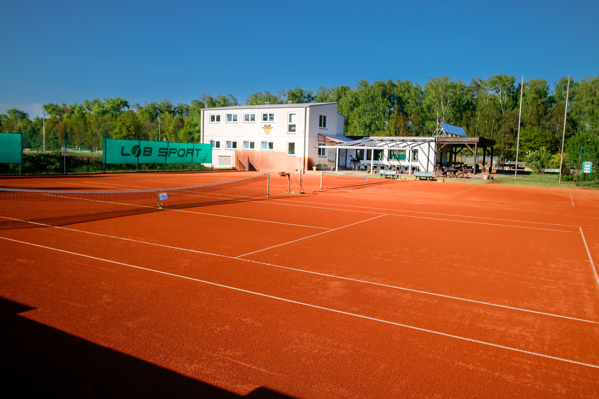 Leerer Tennisplatz aus rotem Sand mit Netz, dahinter ein Gebäude und Bäume.