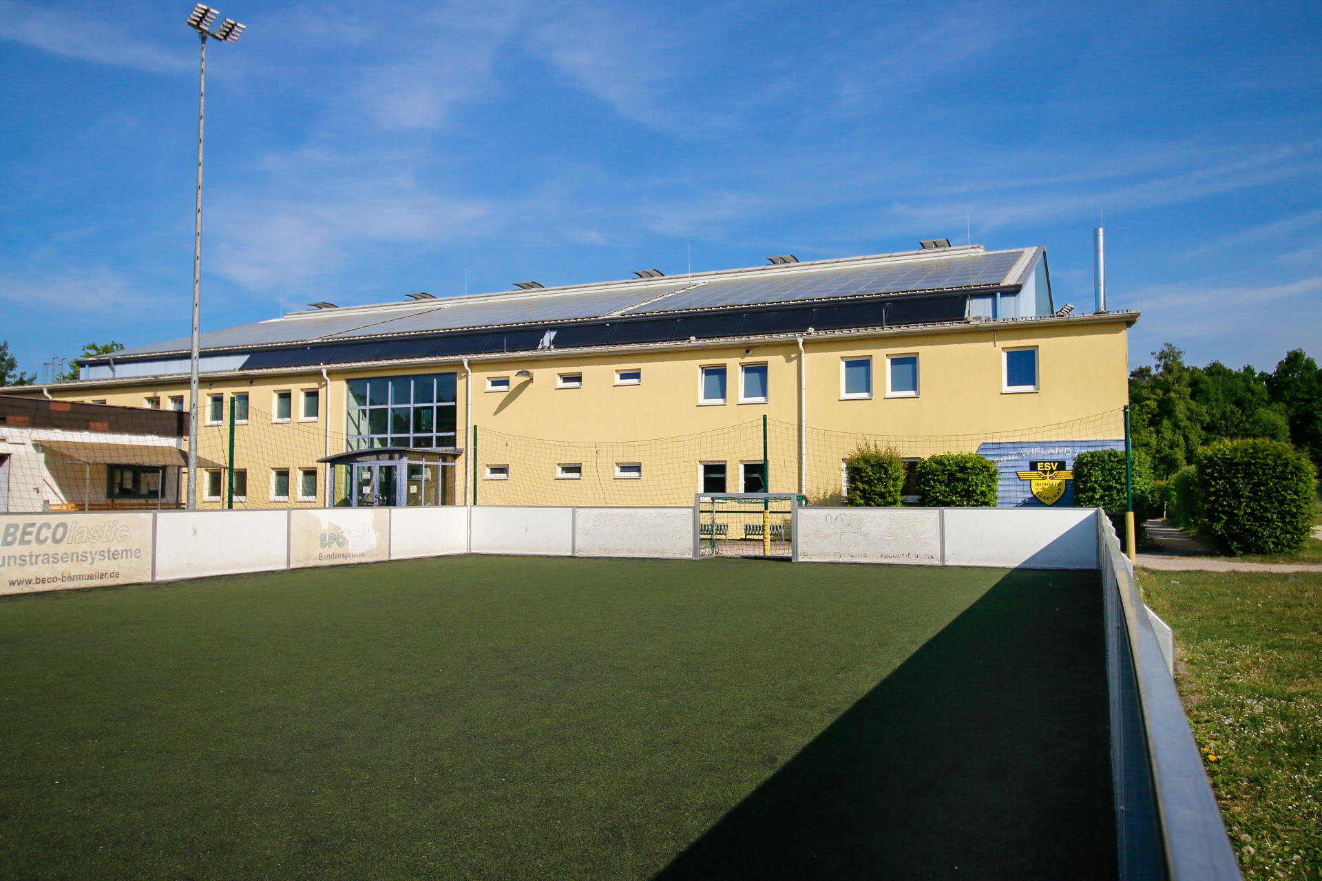 Gelbes Gebäude mit großen Fenstern und Solarpanelen auf dem Dach, davor ein umzäunter Kunstrasenplatz mit Tor. Links eine Flutlichtanlage, im Hintergrund Bäume und blauer Himmel.