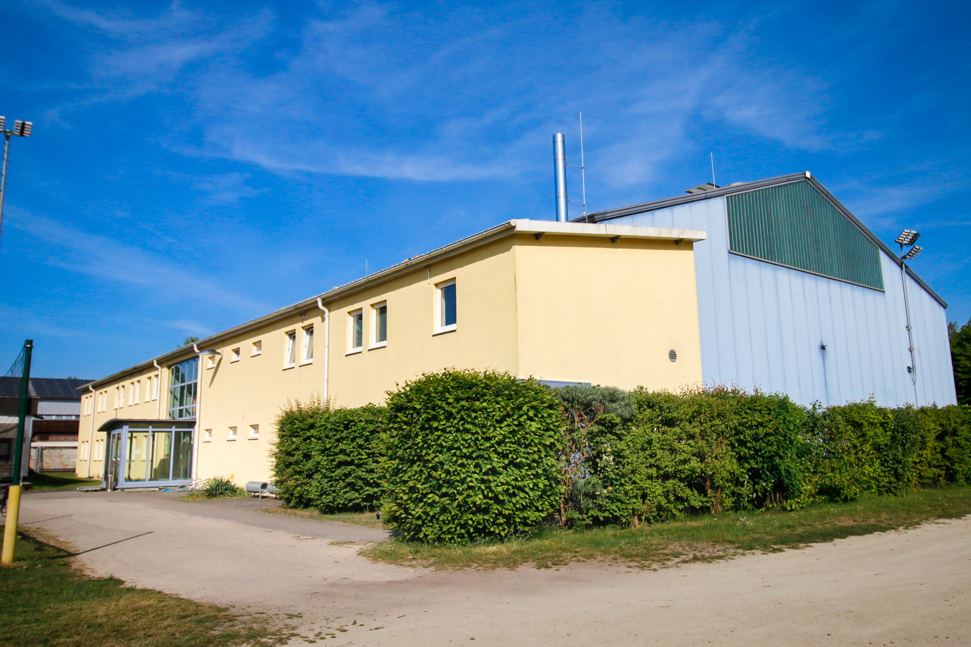 Großes gelbes Gebäude mit mehreren rechteckigen Fenstern und einem verglasten Eingangsbereich, seitlich angrenzend eine große Halle mit grauer Blechfassade und grünem Giebelfeld. Davor ein Schotterweg und grüne Büsche, im Hintergrund blauer Himmel.