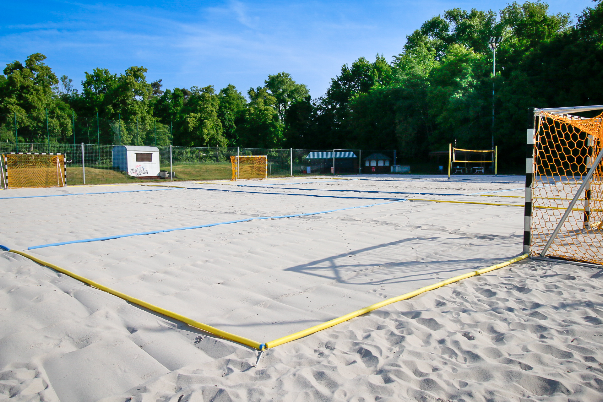 Leeres Beachfeld mit Sand, Spielfeldmarkierungen und mehreren Toren, im Hintergrund Bäume.