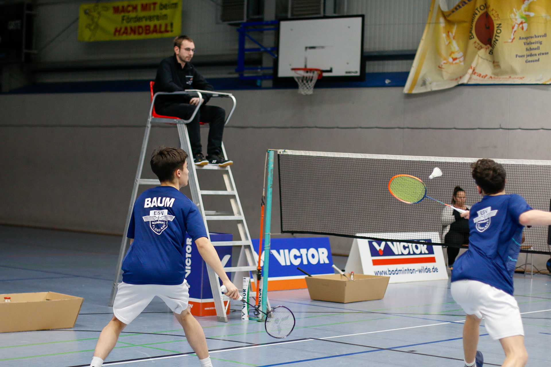 Zwei Badmintonspieler in blauen Trikots des ESV Nürnberg-Flügelrad spielen ein Doppelmatch in einer Sporthalle. Der Spieler links trägt den Namen „BAUM“ auf dem Rücken. Ein Schiedsrichter sitzt erhöht auf einem Stuhl am Spielfeldrand, während ein Shuttlecock über das Netz fliegt. Im Hintergrund sind Banner, eine Basketballkorbanlage sowie Werbetafeln zu sehen.