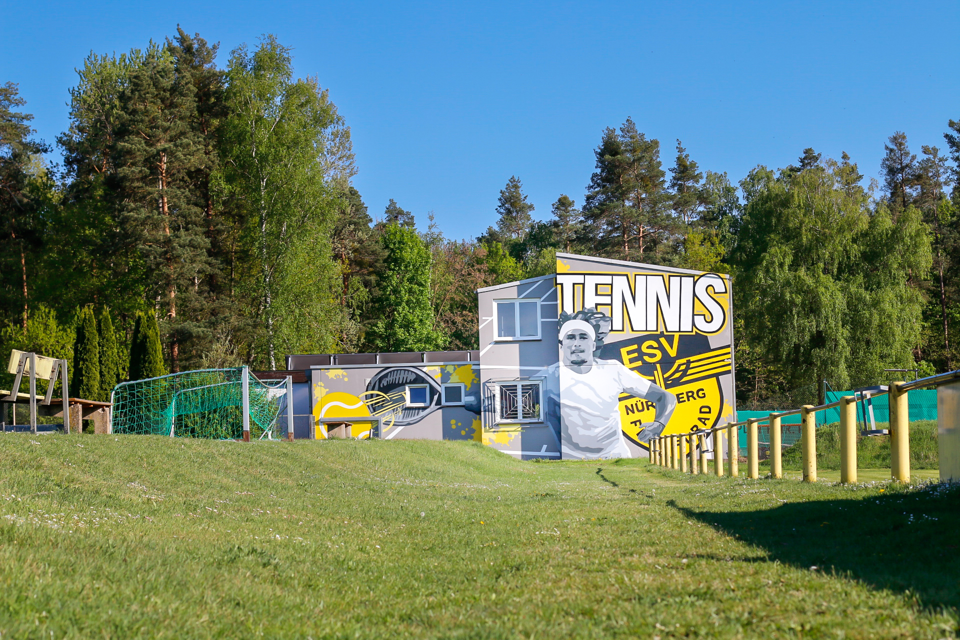 Grünes Sportgelände mit Rasenfläche, im Hintergrund ein graues Gebäude mit großem Wandbild. Darauf ein gezeichneter Tennisspieler in Weiß, daneben die Schrift ‚TENNIS ESV Nürnberg Flügelrad‘ in Schwarz und Gelb. Umgeben von hohen Bäumen und blauem Himmel.