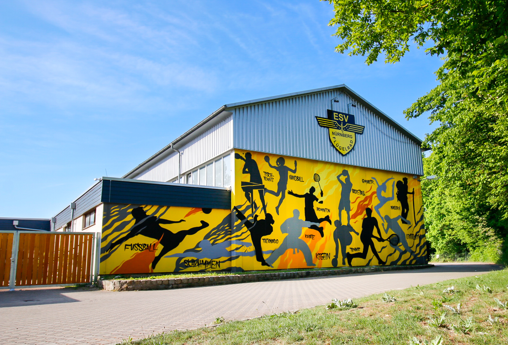 Große Sporthalle mit grauer Metallfassade, an der Stirnseite mit einem gelb-schwarzen Vereinslogo ‚ESV Nürnberg Rangierbahnhof Flügelrad‘. Die Seitenwand ist bunt bemalt mit Silhouetten von Sportlern in Aktion und Schriftzügen wie Fußball, Schwimmen, Tischtennis, Handball, Badminton, Taekwondo, Karate, Kegeln, Tennis, Gymnastik, Basketball, Wandern und Leichtathletik.