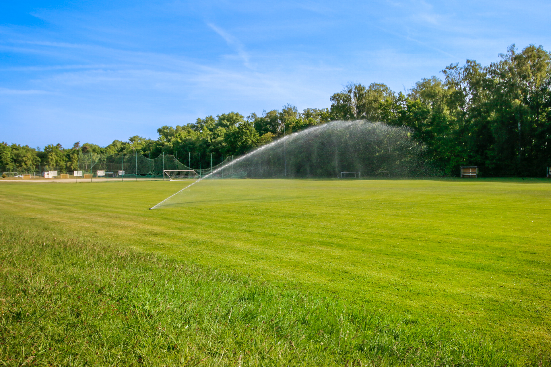 Ein gepflegter, grüner Sportplatz wird mit einer großen Sprinkleranlage bewässert. Im Hintergrund sind Tore, Bäume und ein klarer blauer Himmel zu sehen.