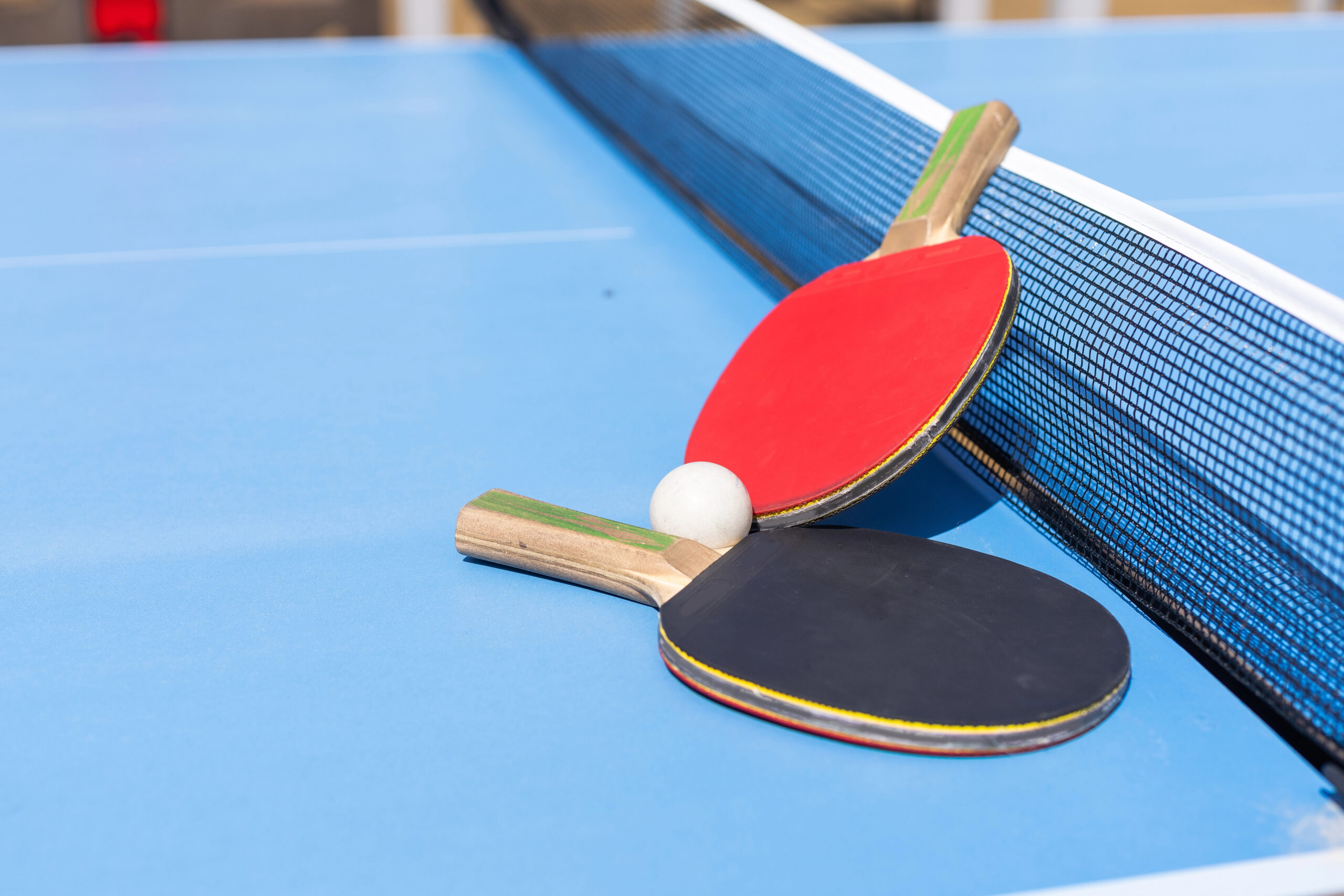 Zwei Tischtennisschläger, einer mit roter und einer mit schwarzer Schlagfläche, liegen über Kreuz auf einer blauen Tischtennisplatte neben einem weißen Ball und dem Netz.