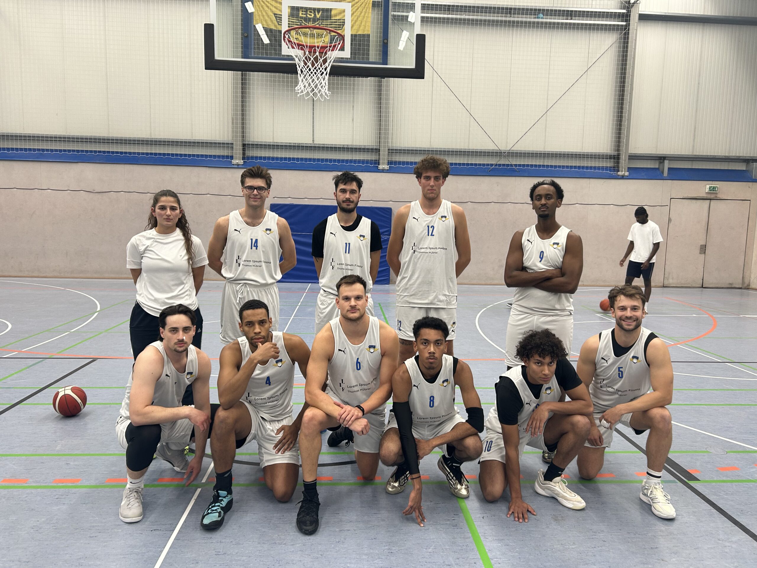Eine Basketballmannschaft posiert in einer Sporthalle für ein Teamfoto. Elf Personen stehen oder knien in zwei Reihen: Die hintere Reihe steht aufrecht, die vordere kniet oder hockt. Alle Spieler tragen weiße Basketballtrikots mit blauen Nummern und einem Vereinslogo, während eine Person in der hinteren Reihe ein weißes Shirt trägt. Hinter dem Team hängt ein Basketballkorb an der Wand. Der Hallenboden zeigt bunte Linienmarkierungen, und rechts im Hintergrund ist eine weitere Person sichtbar. Die Stimmung wirkt konzentriert und teamorientiert.