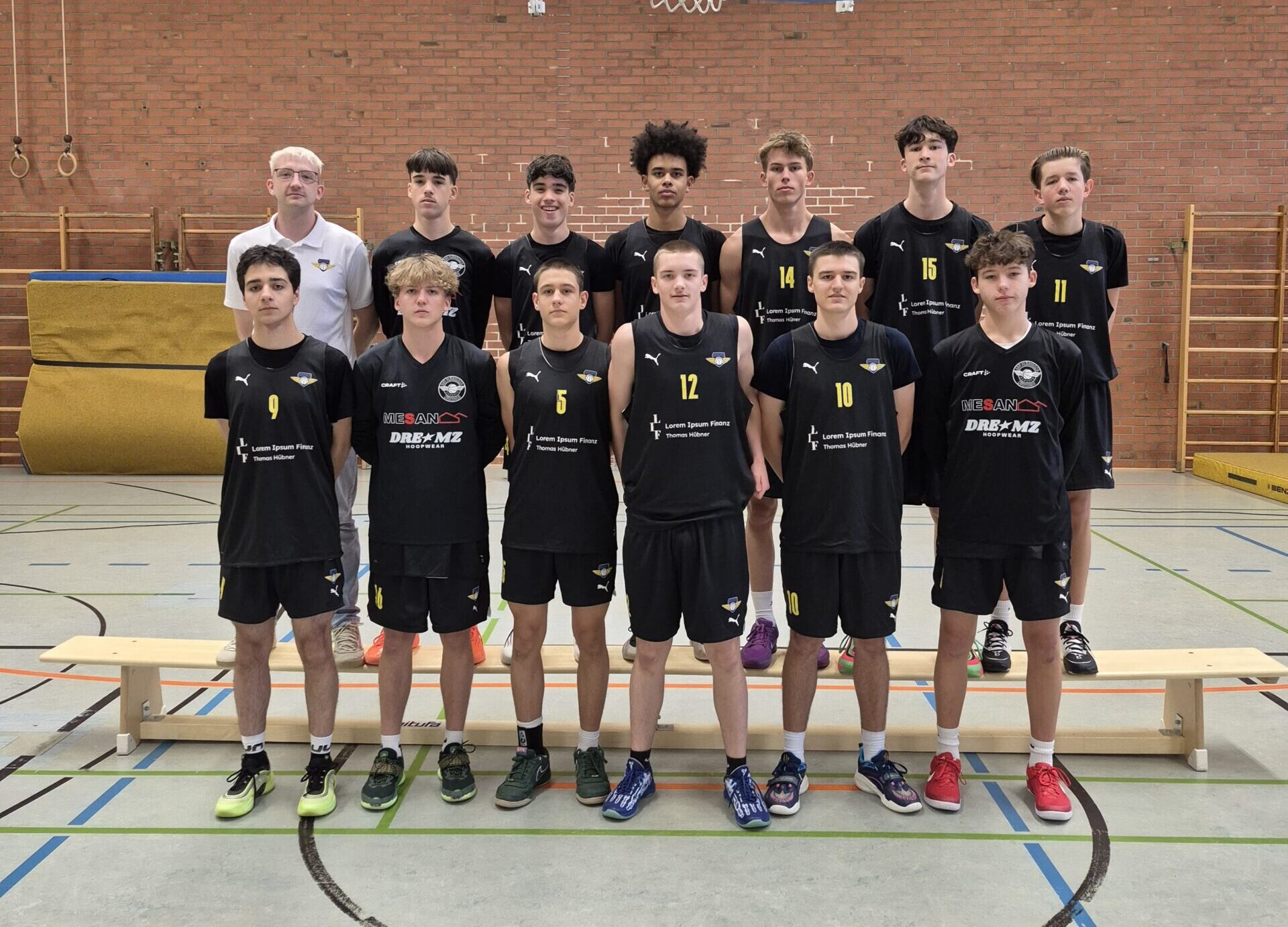 Eine Jugend-Basketballmannschaft posiert in einer Sporthalle für ein Teamfoto. Vierzehn Spieler und ein Coach stehen in zwei Reihen unter einem Basketballkorb. Alle Spieler tragen schwarze Trikots mit gelben Nummern und Sponsorenlogos, während der Coach ein weißes Shirt trägt. Die Gruppe steht auf und vor einer Holzbank, im Hintergrund sind Turnhallenwände aus rotem Backstein, Matten und Sportgeräte zu sehen. Die Stimmung wirkt geordnet und konzentriert.