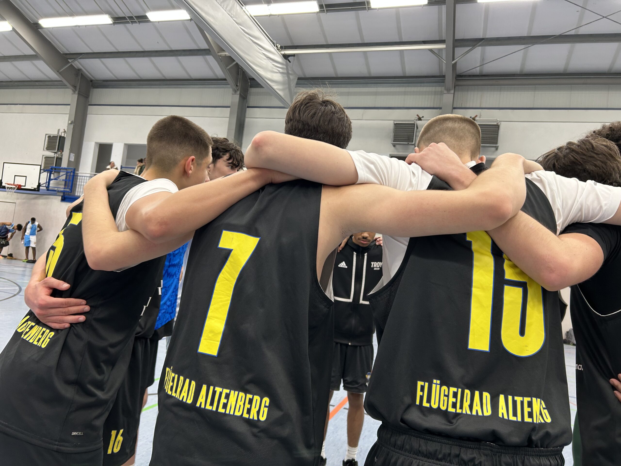 Mehrere junge Basketballspieler stehen dicht beieinander in einem Mannschaftskreis und legen die Arme über die Schultern der Teamkollegen. Sie tragen schwarze Trikots mit großen gelben Rückennummern und dem Aufdruck „Flügelrad Altenberg“. Die Szene spielt in einer Sporthalle; im Hintergrund sind Basketballkörbe, Tribünen und weitere Personen zu sehen. Die Stimmung wirkt konzentriert und gemeinschaftlich, als ob das Team sich kurz vor dem Spiel oder während einer Auszeit motiviert.
