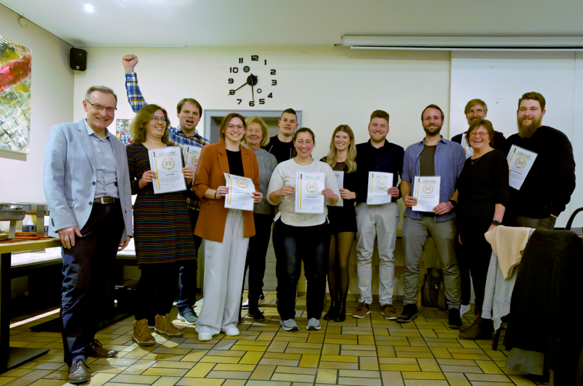 Gruppenfoto von etwa zwölf Personen in einem hellen Raum, die gemeinsam in die Kamera lächeln. Mehrere von ihnen halten Urkunden mit der Aufschrift „25“ in den Händen, vermutlich zur Ehrung eines Jubiläums. Eine Person im Hintergrund hebt jubelnd den Arm. Im Raum sind ein Wandbild, eine Uhr und ein ausgerollter Beamer zu sehen.