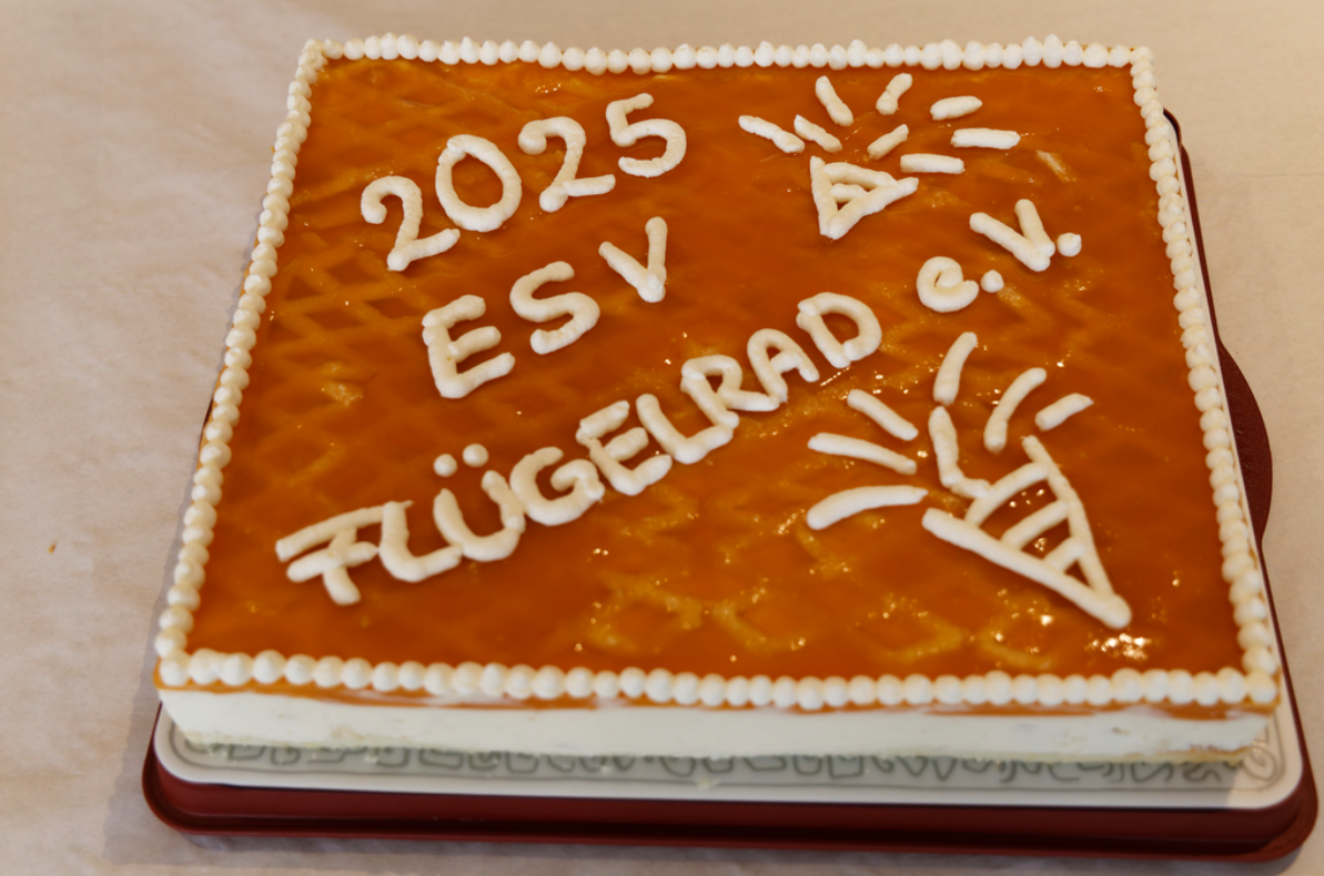 Rechteckige Torte mit orangefarbener Glasur und weißer Zuckerschrift „2025 ESV Flügelrad e.V.“ sowie dekorativen Symbolen; am Rand ist sie mit kleinen weißen Tupfen verziert und steht auf einer Servierplatte.